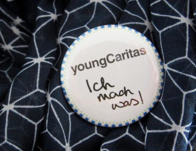 Ein Anstecker mit dem Schriftzug 'youngCaritas' und der handschriftlichen Aufschrift 'Ich mach was!'. Er liegt auf einem Stoff mit geometrischem Muster.