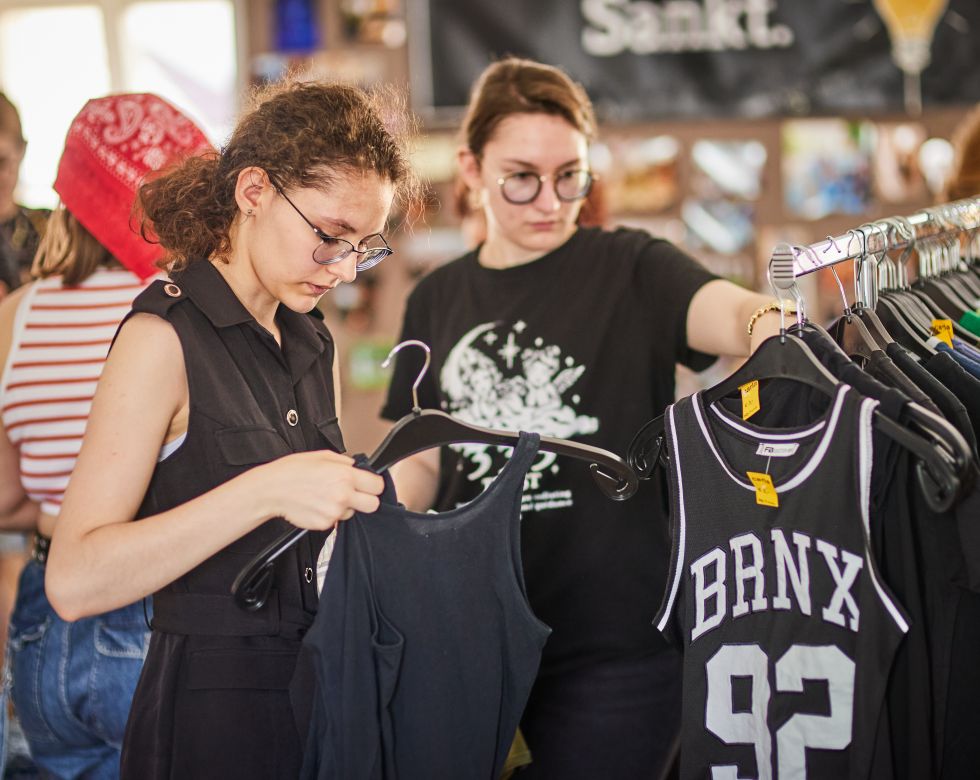 Junge Frauen stöbern in einem Second-Hand-Laden und halten verschiedene Kleidungsstücke in der Hand.