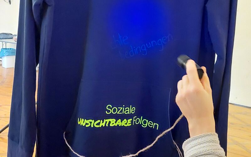 Ein schwarzer Hoodie, auf den mit einem Laserpointer Worte projiziert werden, darunter "Soziale Unsichtbare Folgen".