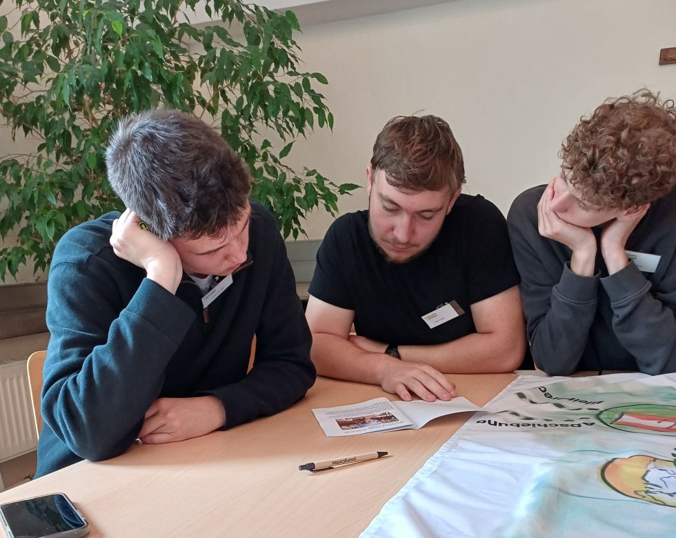 Drei junge Männer sitzen an einem Tisch und lesen konzentriert in einem Heft während einer besinnlichen Diskussion.