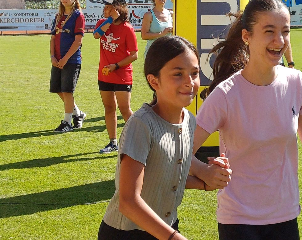 Zwei Mädchen lachen und halten sich beim Laufen an den Händen fest. Im Hintergrund sind andere Kinder und Erwachsene beim Training auf dem Rasen.