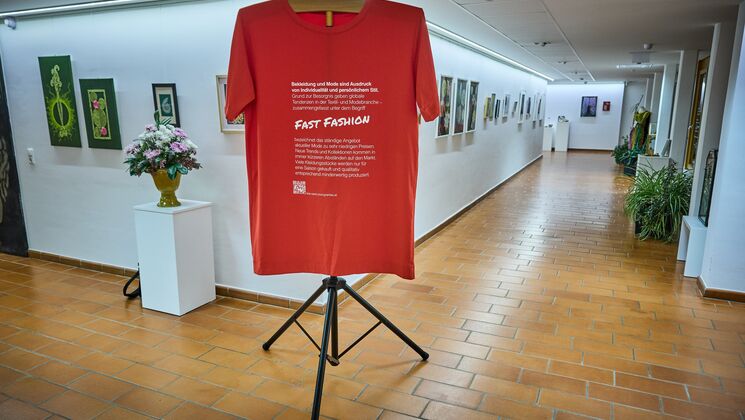 Ein Ausstellungsstück zu Fast Fashion. Es ist ein riesiges rotes T-Shirt mit weißem Text zu sehen.