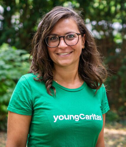Junge Frau mit grünem T-Shirt, auf dem ein youngCaritas Schriftzug ist, posiert vor grünem Naturhintergrund.