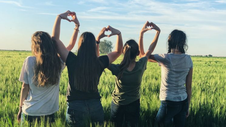 Fünf Frauen stehen in einem Feld und formen mit ihren Händen Herzen. Die Sonne scheint und der Himmel ist blau.