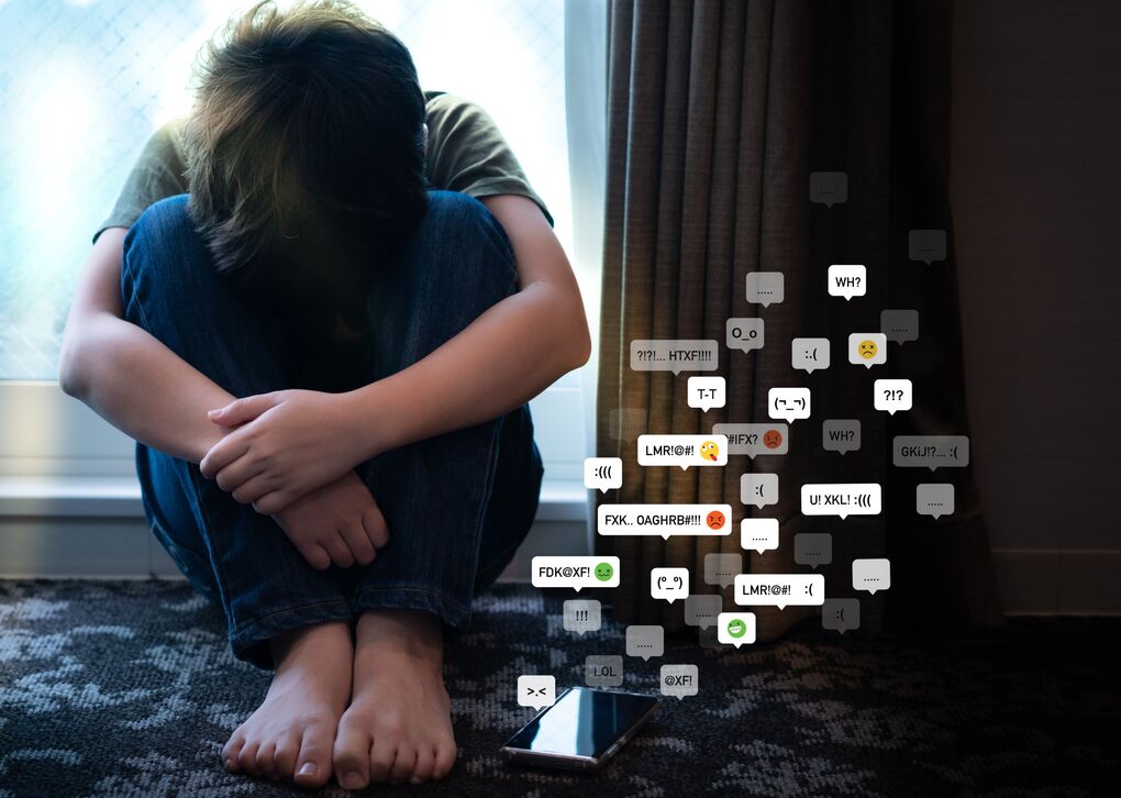 Ein Junge sitzt auf dem Boden, umgeben von negativen Nachrichten und Emojis auf dem Bildschirm, die seine Traurigkeit und Isolation widerspiegeln.