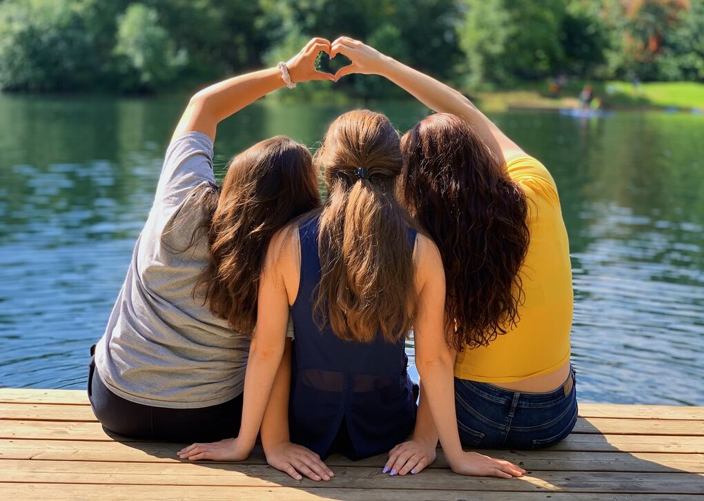 Drei Frauen sitzen auf einem Holzsteg am Wasser und bilden mit ihren Händen ein Herz.