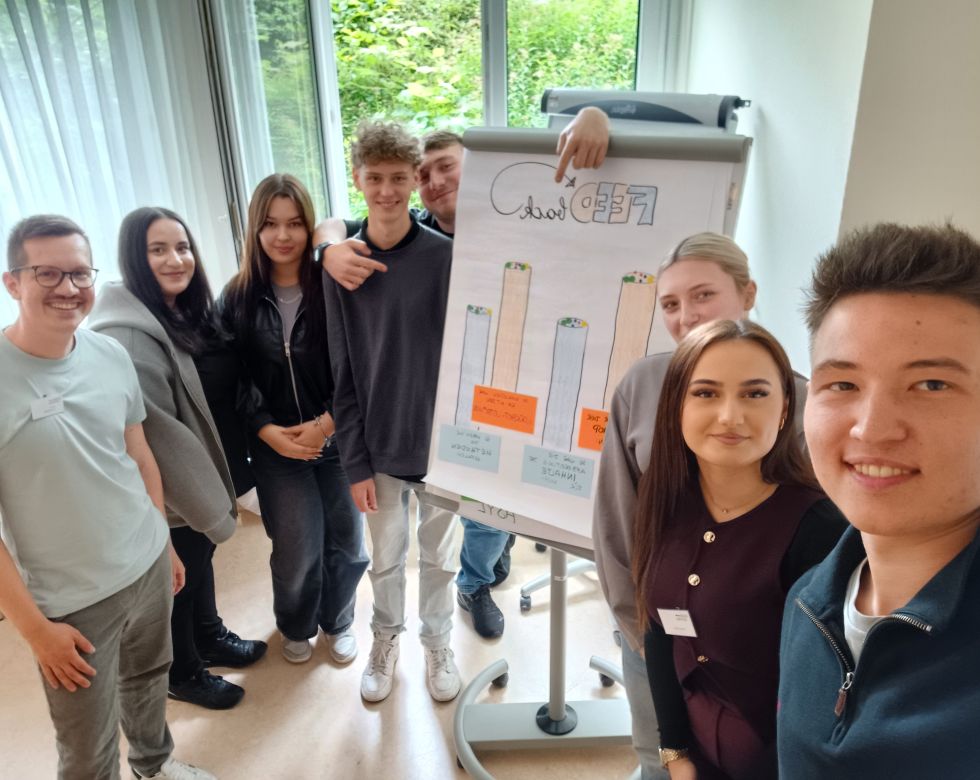 Gruppenfoto von Teilnehmern in einer Besprechung mit einem Flipchart im Hintergrund, auf dem Ergebnisse präsentiert werden.