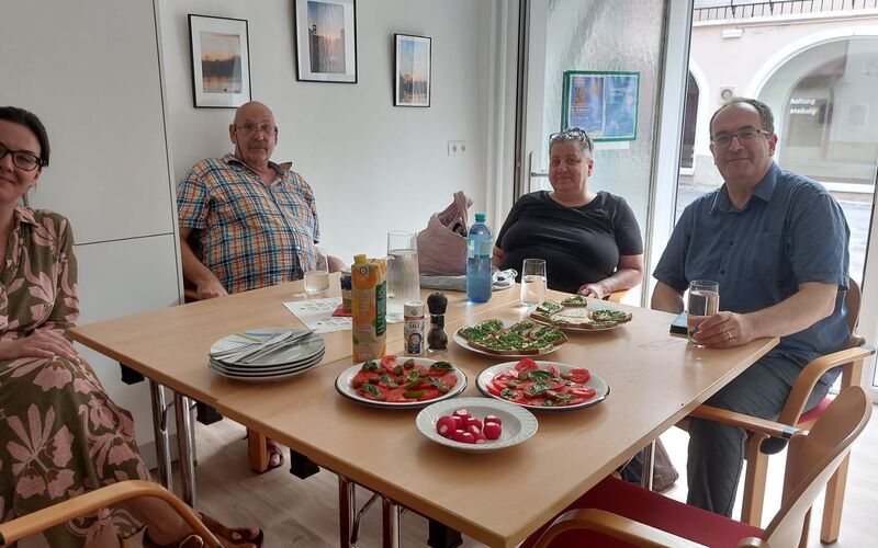 Menschen sitzen um eine Tisch, der gedeckt ist mit Tellern mit Gemüse und Broten und Getränken