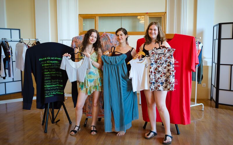 Drei junge Frauen posieren mit Kleidungsstücken in der Hand vor der Ausstellung "tragbar?"
