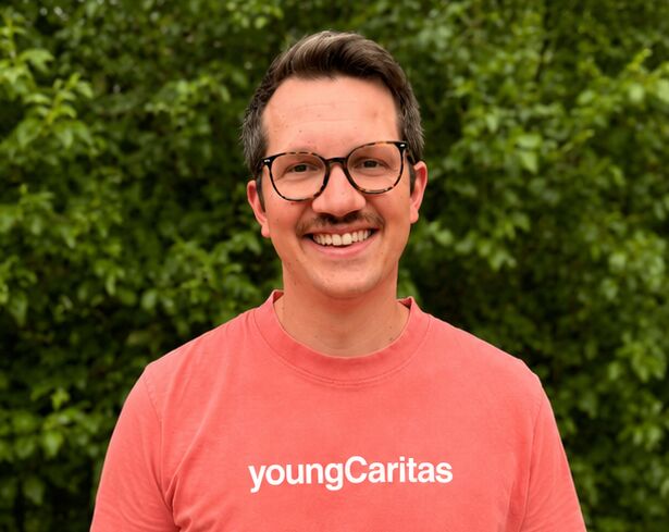 Ein junger Mann trägt ein lachsfarbenes T-Shirt mit der Aufschrift youngCaritas und lächelt