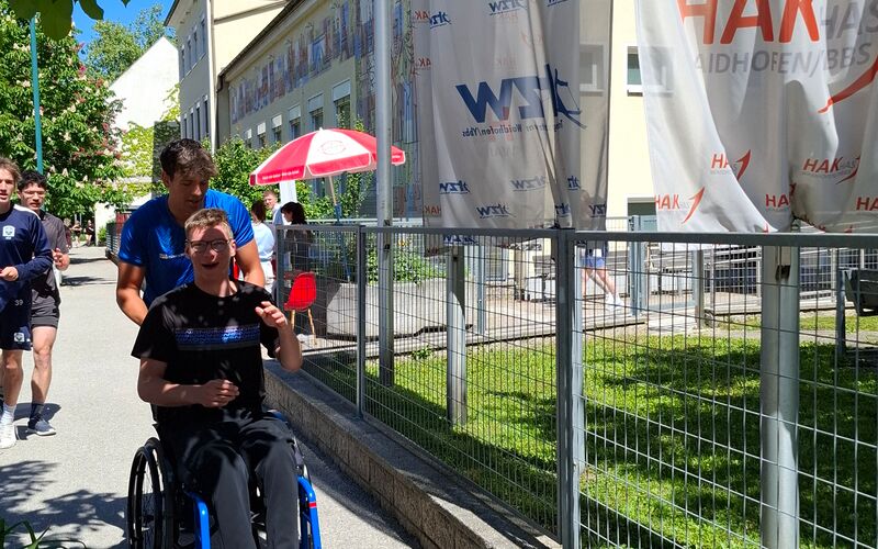 Ein junger Mann im Rollstuhl wird von einem Sportler am Gehsteig angeschoben. Daneben ist ein Zaun und Banner vor einer Schule.