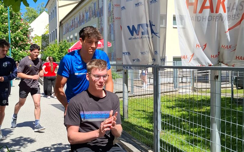 Ein junger Mann im Rollstuhl wird von einem Sportler am Gehsteig angeschoben. Daneben ist ein Zaun und Banner vor einer Schule.