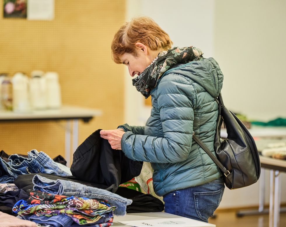 Eine Frau blickt auf eine schwarze Jacke, während sie Kleidung auf einem Tisch durchstöbert.