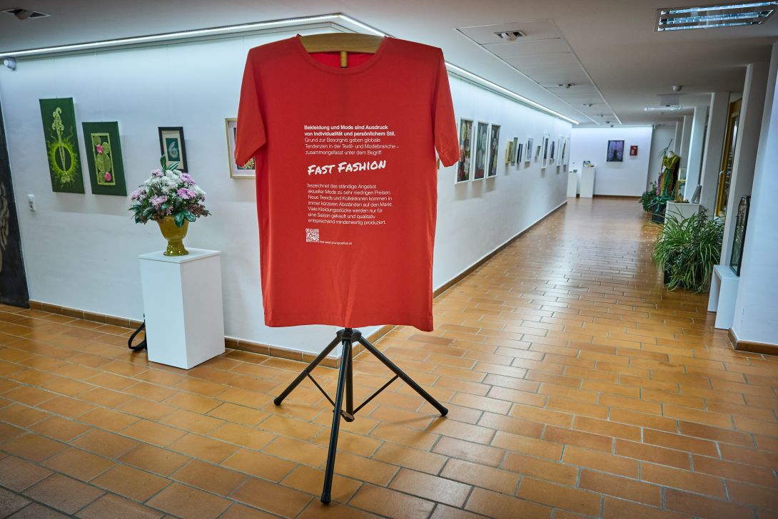 Ein rotes T-Shirt mit dem Schriftzug 'Fast Fashion' in einer Galerie, umgeben von Kunstwerken an den Wänden.