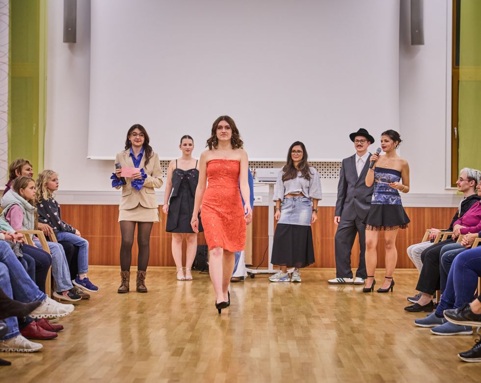 Eine Gruppe junger Models präsentiert verschiedene Outfits bei einer Modenschau. Zuschauer sitzen im Publikum und beobachten aufmerksam.